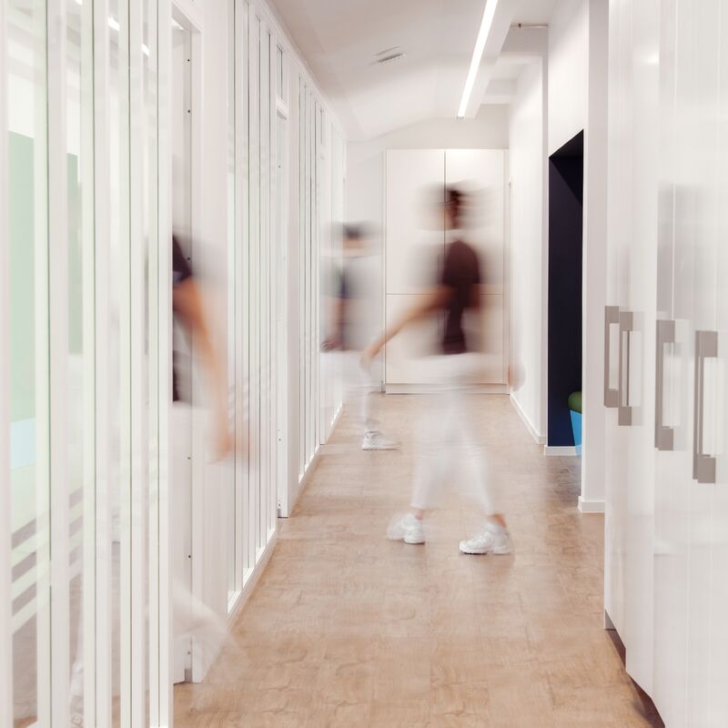 Mehrere Personen in weißer Kleidung bewegen sich durch einen hellen Büroflur, aufgenommen in Bewegungsunschärfe.