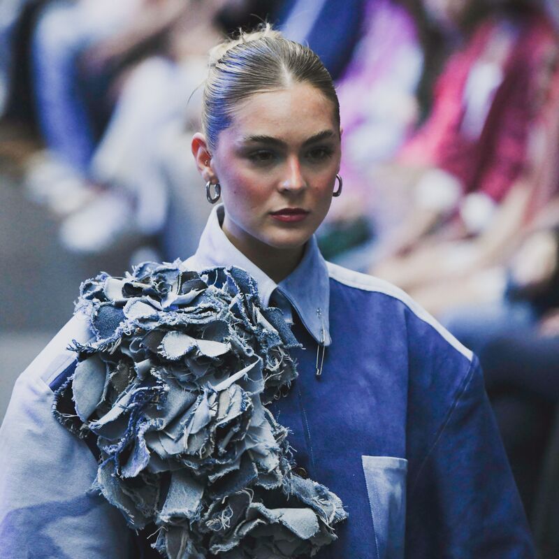 Ein Model läuft über den Laufsteg und trägt ein blaues, kunstvoll gestaltetes Jeans-Outfit mit großen, blumenartigen Applikationen aus Denim auf der Schulter. Der Hintergrund ist unscharf und zeigt Zuschauer am Rand der Modenschau.