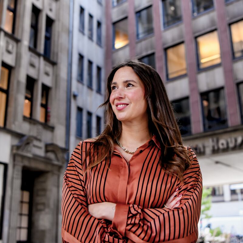 Eine Frau in einer gestreiften, rostfarbenen Bluse steht mit verschränkten Armen lächelnd in einer Altstadtgasse mit modernen Bürogebäuden im Hintergrund.